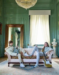 Two musicians holding electric guitars seated on a vintage floral sofa in a green paneled room photographed by Elizabeth Lavin