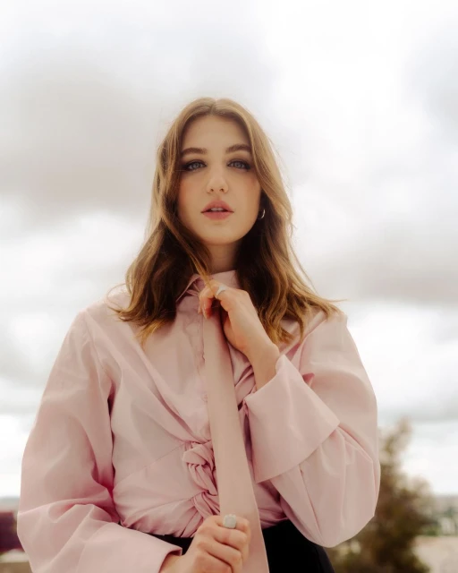 Portrait of a young woman in a pink blouse with a soft tie, posing outdoors against a cloudy sky with a neutral facial expression.