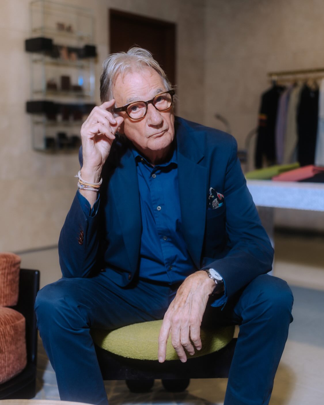 Elderly man in a blue suit sits indoors, wearing glasses, with a neutral expression. He is in a naturally lit room with neutral walls.