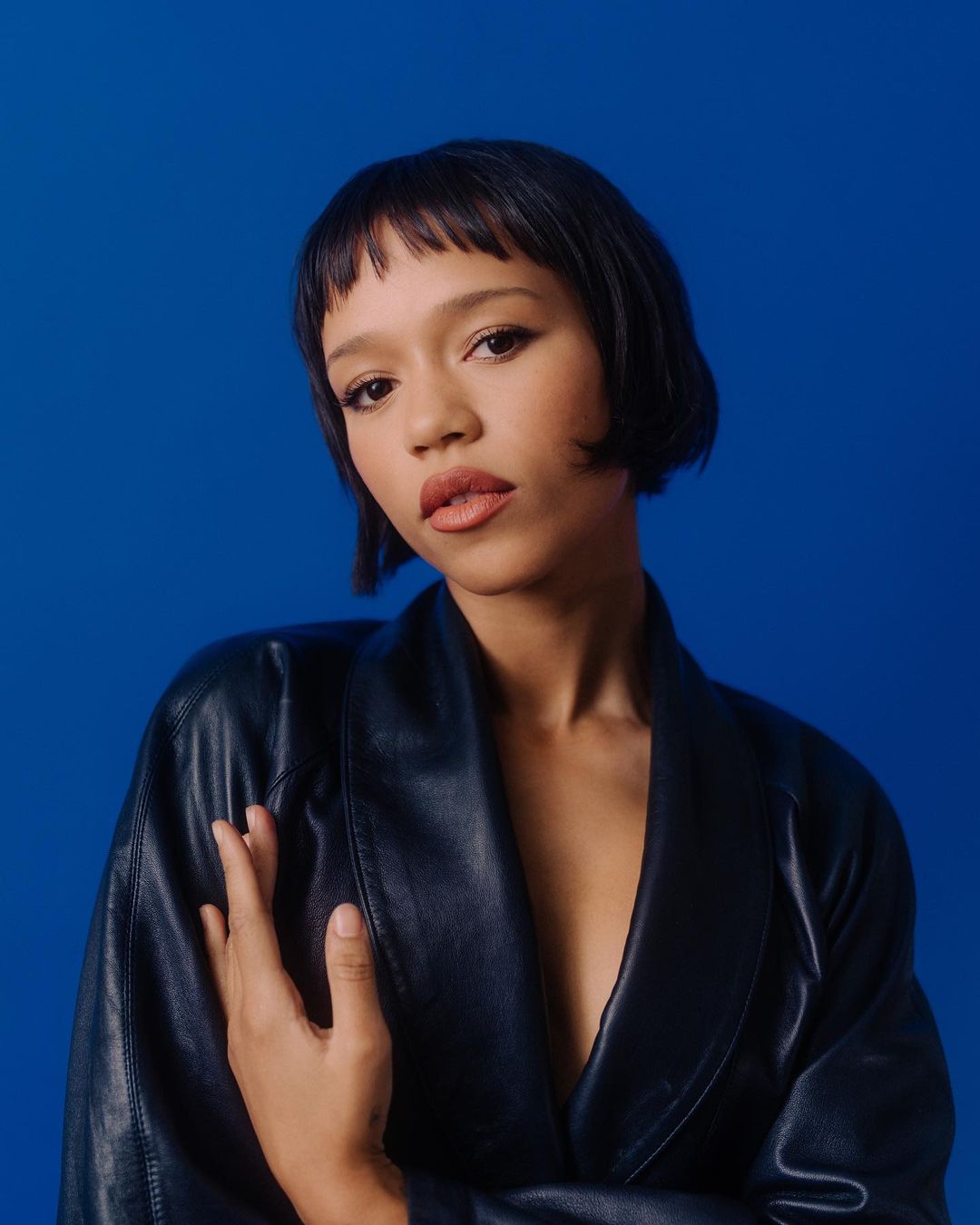 Portrait of a woman with short dark hair and bangs wearing a dark blue leather jacket against a bright blue studio backdrop.