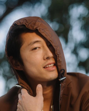 Portrait of an Asian man wearing a brown hooded jacket with a drawstring outdoors with bokeh foliage background