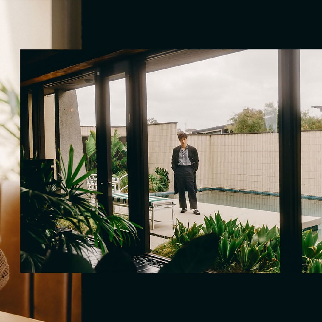 Adult man in a suit stands near a pool outside a home with large glass windows and green plants in the foreground.