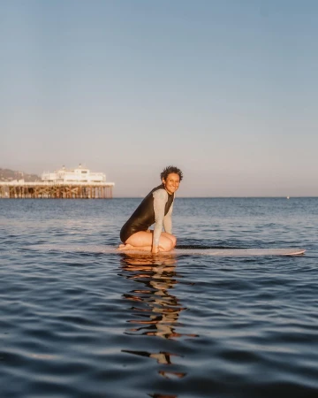 Woman in wetsuit sitting on surfboard in the ocean with pier in background, reflecting in the water, creating a serene beach scene.