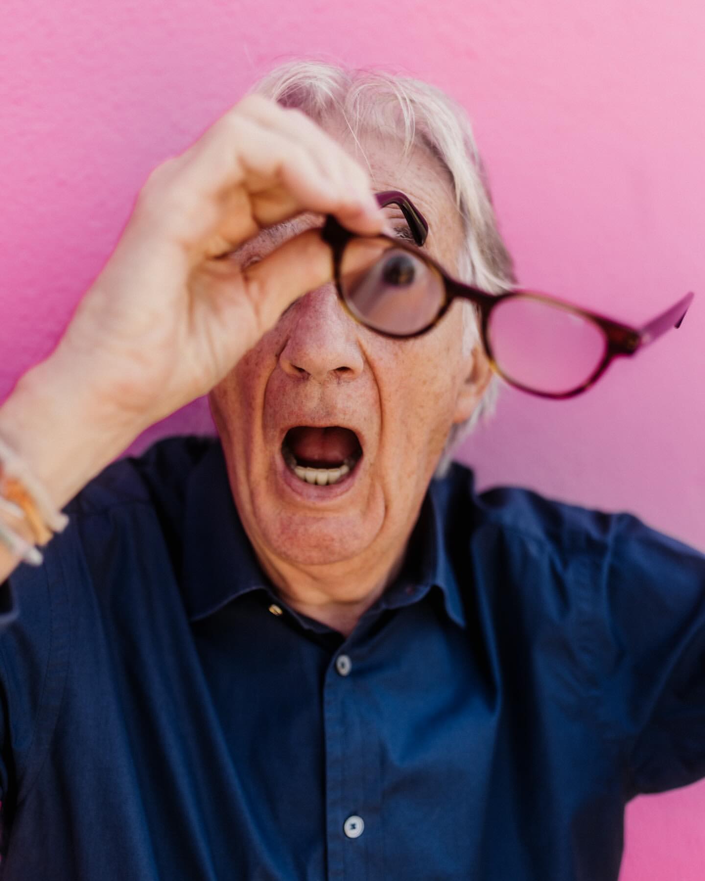 Elderly man with gray hair holds glasses up to one eye with an open mouth expression against a pink background in a portrait.