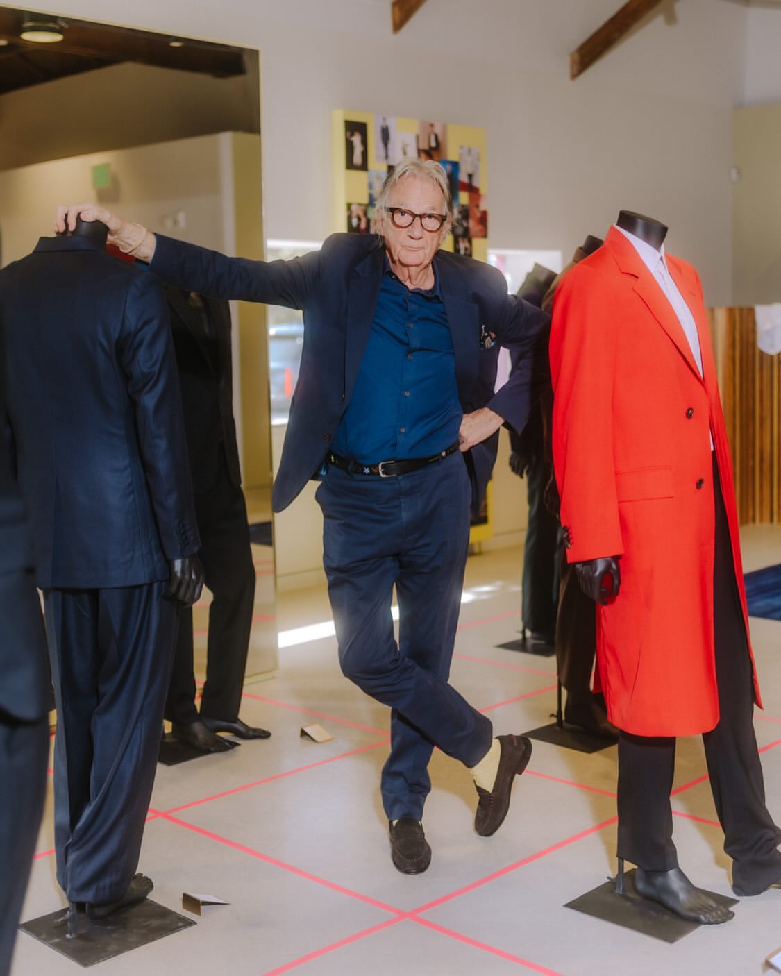 Older man in a blue suit strikes a pose in an indoor space surrounded by mannequins in modern coats and suits on a tiled floor.