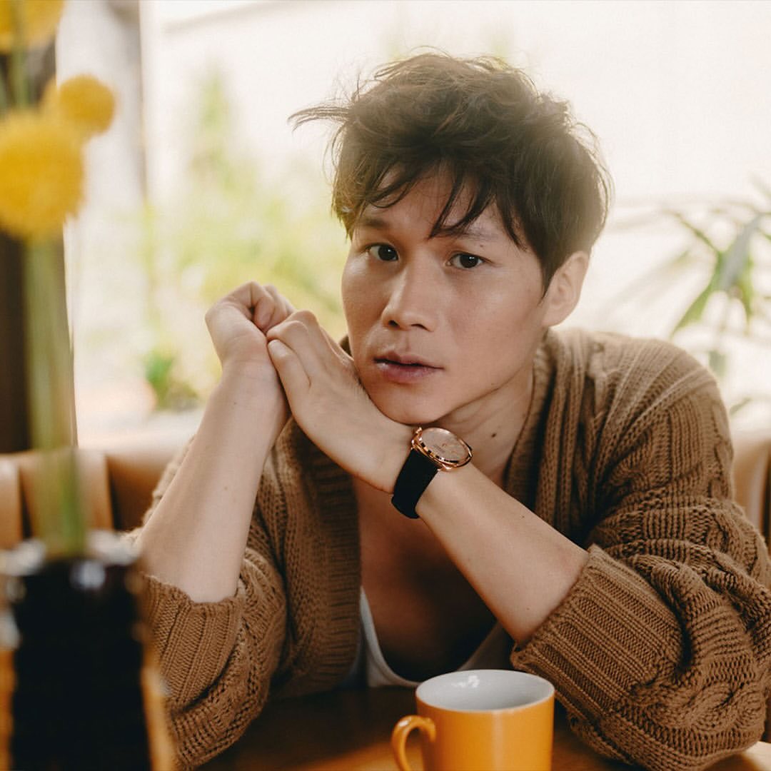 Portrait of a Korean adult male wearing a brown knit cardigan with a cup of coffee in an indoor cafe setting during the daytime.