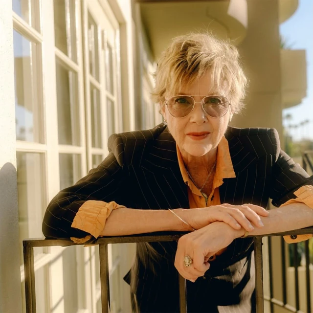Elegant portrait of an older woman wearing aviator sunglasses and a pinstripe blazer, captured in natural light on a balcony.