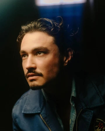 Portrait of a man with dark curly hair and a mustache wearing a blue jacket against a dark background