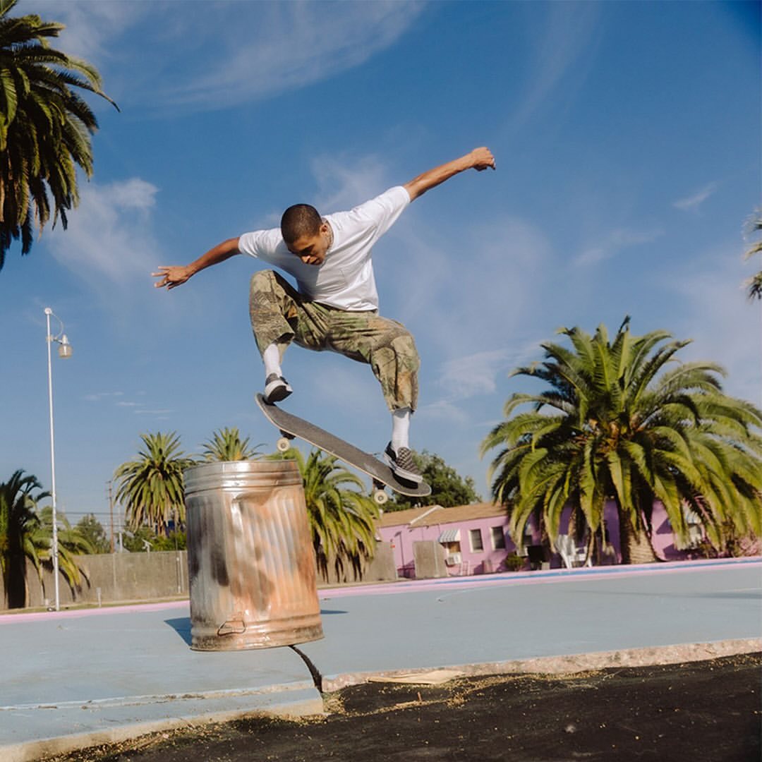 An action shot of a skateboarder performing a trick in mid-air over a trash can, set against a blue sky with palm trees.