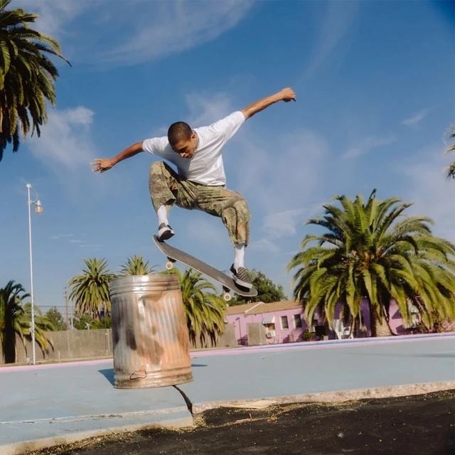 An action shot of a skateboarder performing a trick in mid-air over a trash can, set against a blue sky with palm trees.