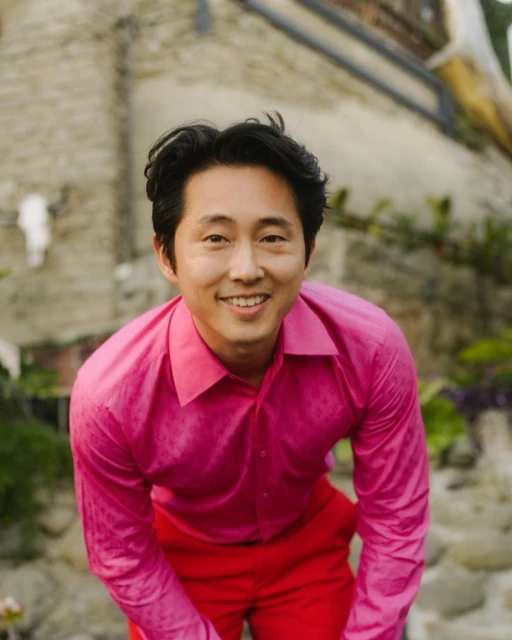 Steven Yeun smiles outdoors in a pink dress shirt and red pants, posing against a textured stone wall with green foliage.