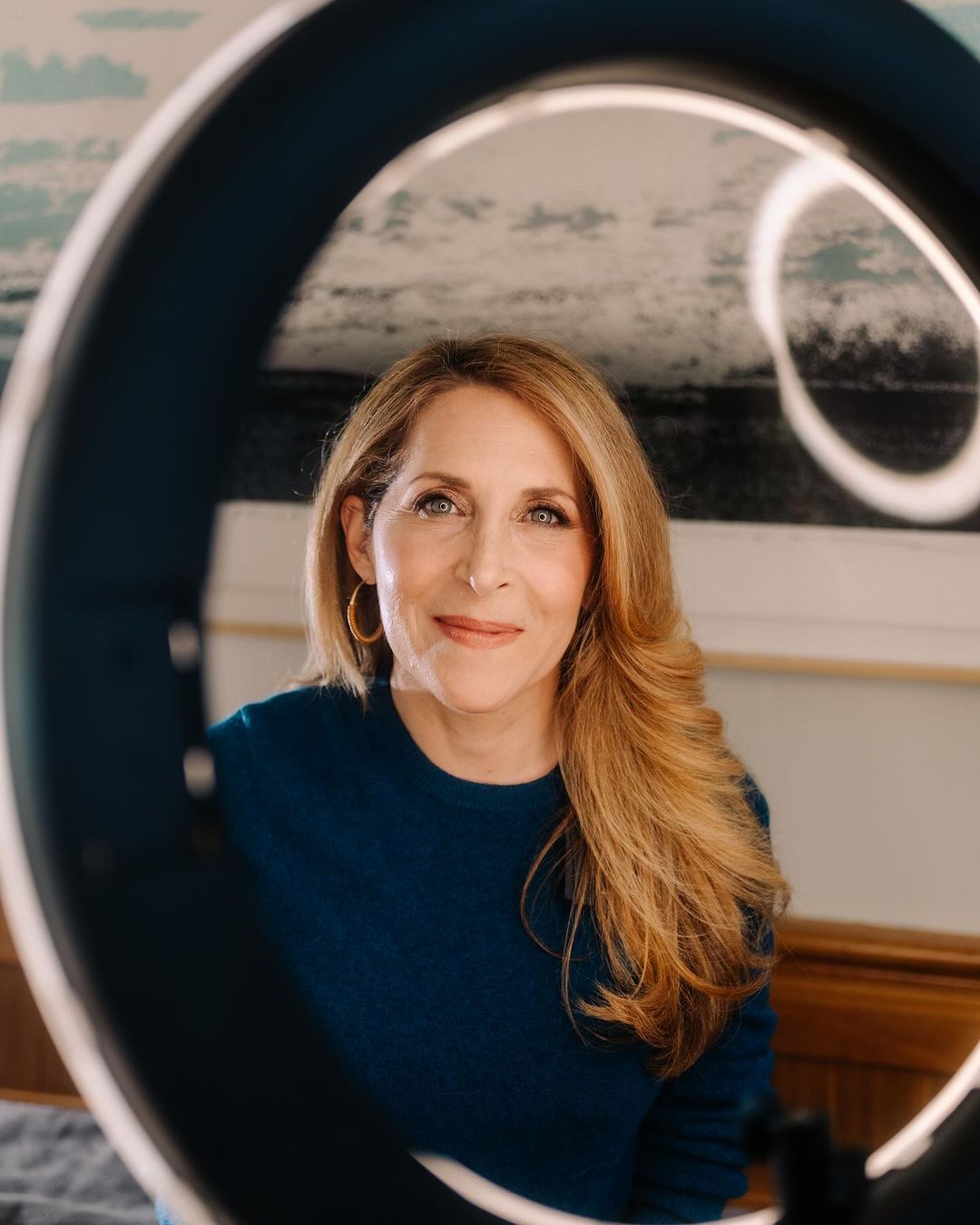 Close-up portrait of a smiling adult woman with long blonde hair, wearing a blue sweater, framed by a ring light indoors.