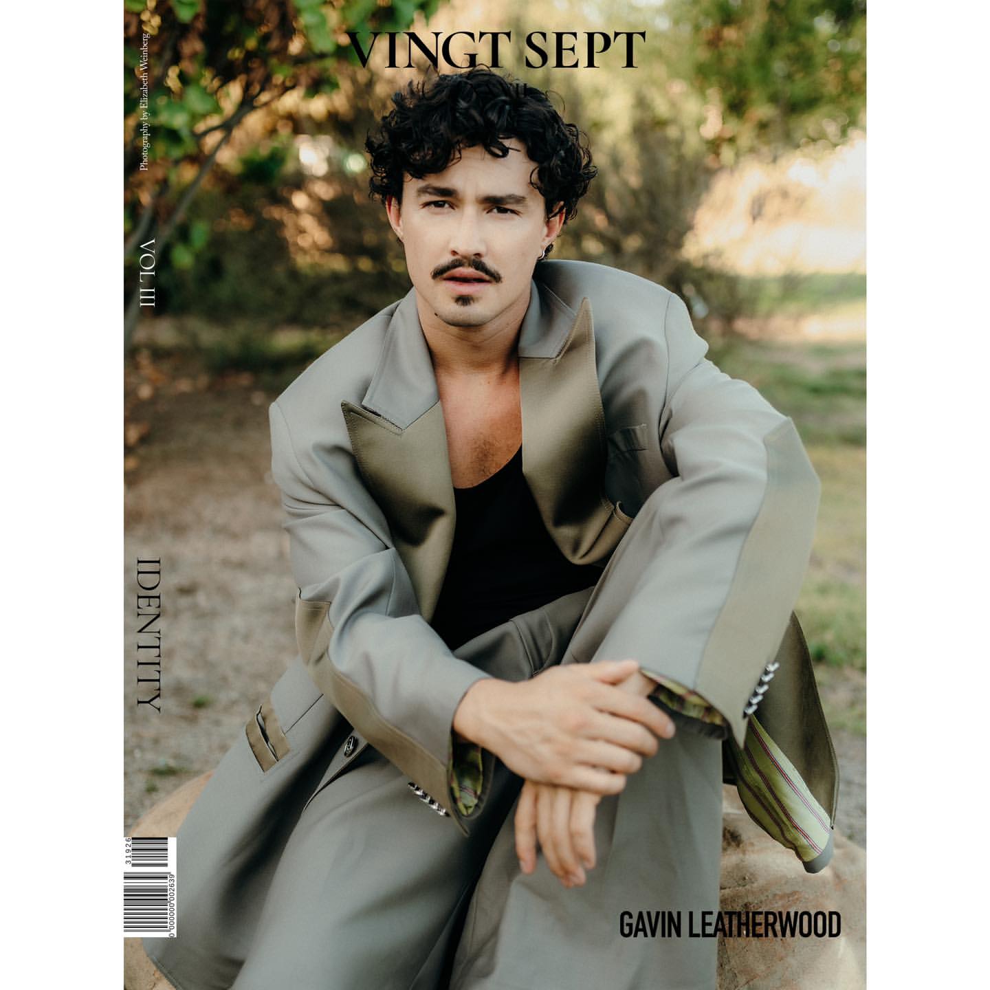 Magazine cover featuring a man in an oversized suit, seated outdoors with a neutral backdrop of trees and natural light.