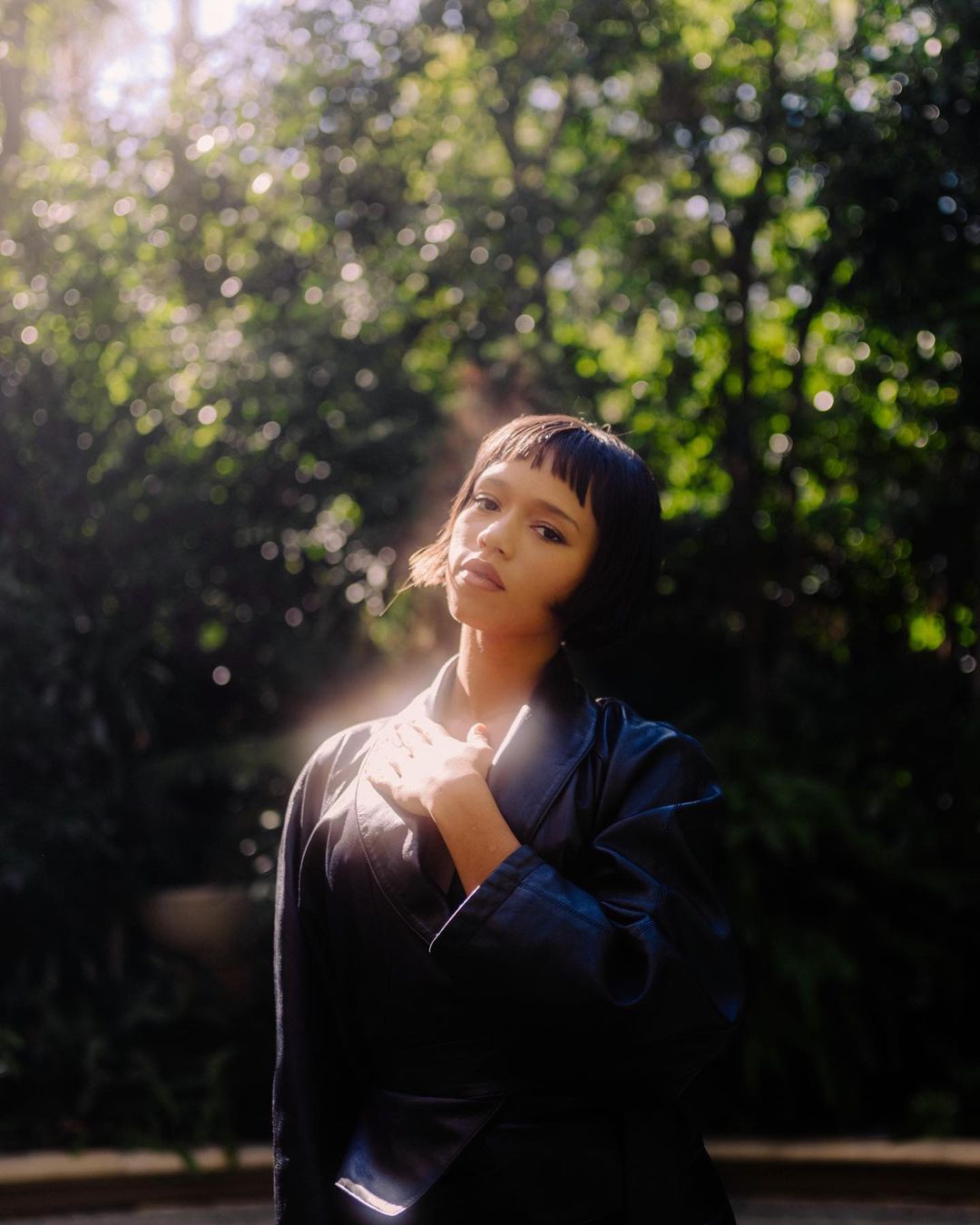 Portrait of a young woman with short dark hair, wearing a black jacket, posing outdoors in a leafy park setting on a sunny day.