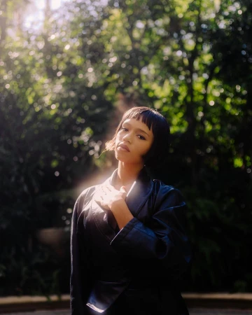 Portrait of a young woman with short dark hair, wearing a black jacket, posing outdoors in a leafy park setting on a sunny day.