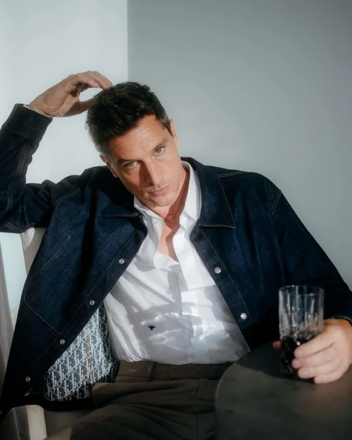 An adult male sits indoors wearing a denim jacket and white shirt, holding a glass. Gray wall background with a table and natural light.