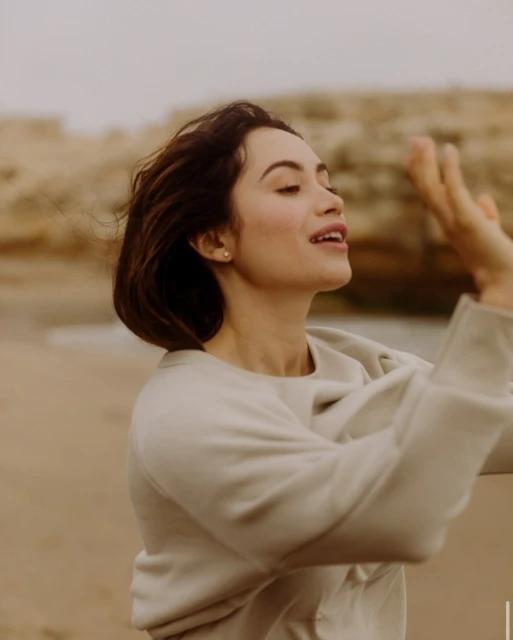 A woman smiles on a beach wearing a light-colored sweatshirt with cliffs in the background. The weather is overcast with diffused lighting.