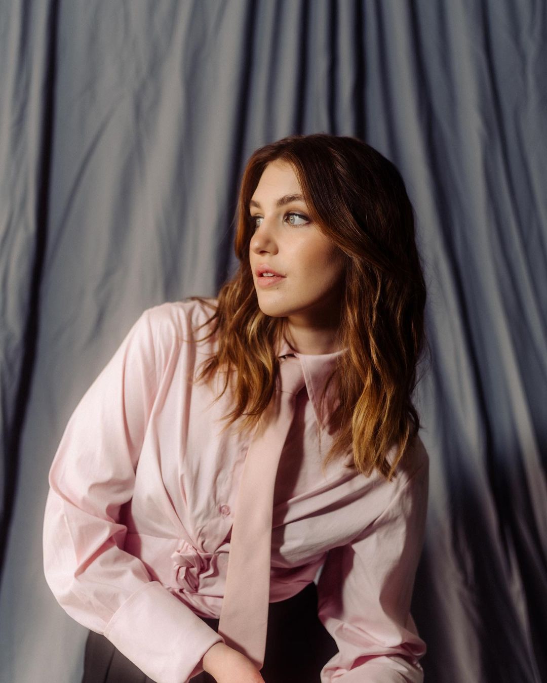 Portrait of a young woman with wavy brown hair wearing a pink button-down shirt and tie in front of a grey draped background.