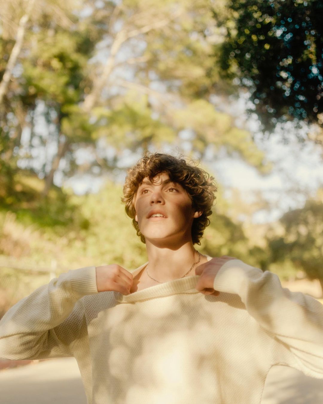 Portrait of a curly-haired young adult male wearing an off-white sweater, looking upward outdoors with trees and sunlight in the background.