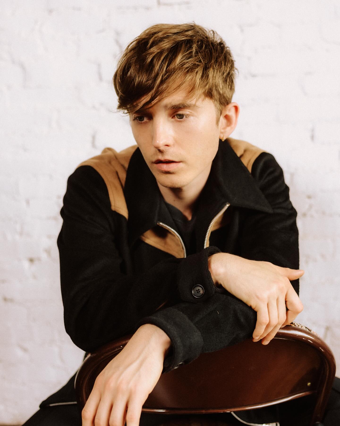 Portrait of a young adult man with brown hair in a black jacket sitting on a brown chair in front of a white brick wall indoors.
