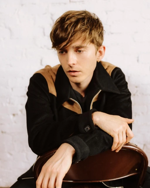 Portrait of a young adult man with brown hair in a black jacket sitting on a brown chair in front of a white brick wall indoors.