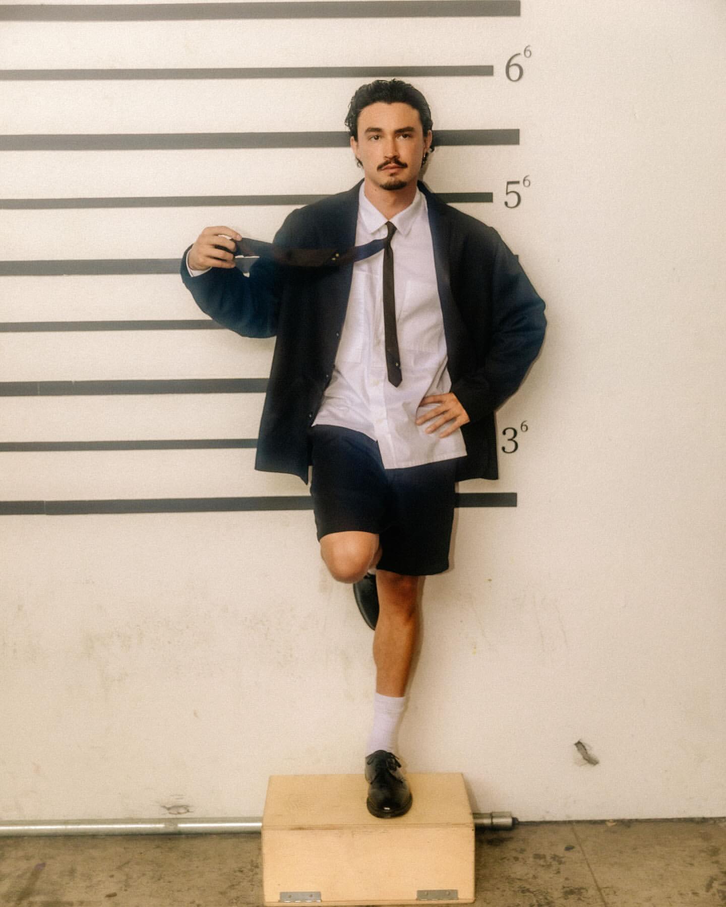 Adult male in a suit jacket, tie, and shorts poses against a height chart on a concrete wall, standing on a small platform.
