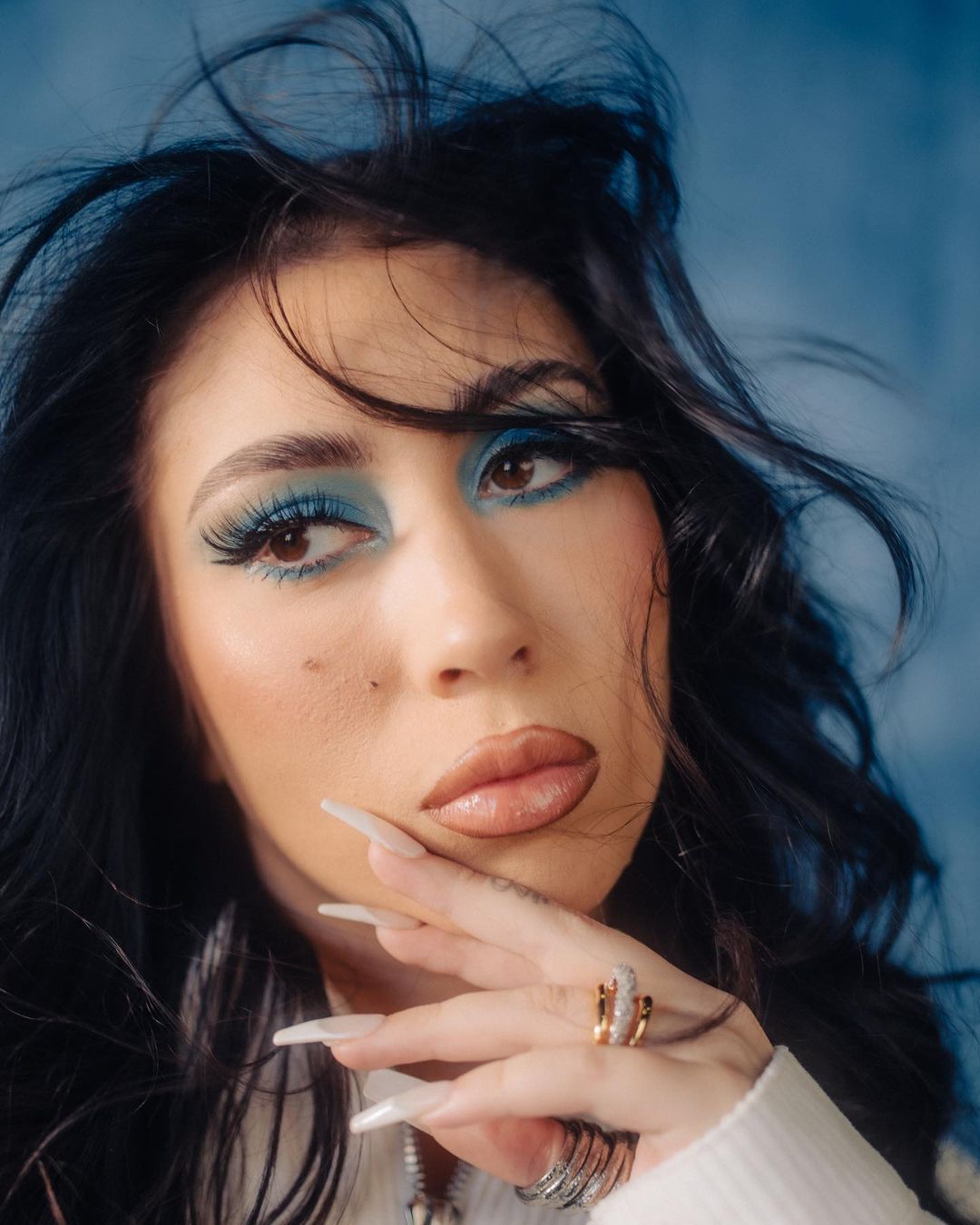 Close-up portrait of a stylish adult female with blue eyeshadow, long nails, and rings against a mottled blue backdrop.