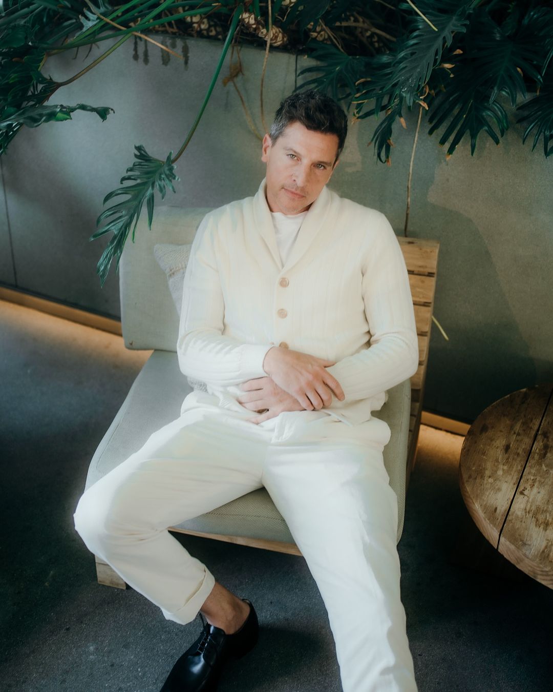 Man in a cream cardigan and matching trousers seated in a chair beneath lush tropical plants, photographed by Elizabeth Weinberg.