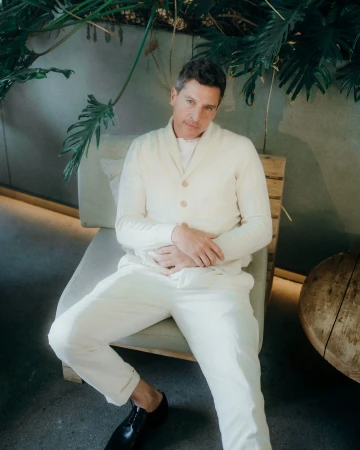 Man in a cream cardigan and matching trousers seated in a chair beneath lush tropical plants, photographed by Elizabeth Weinberg.