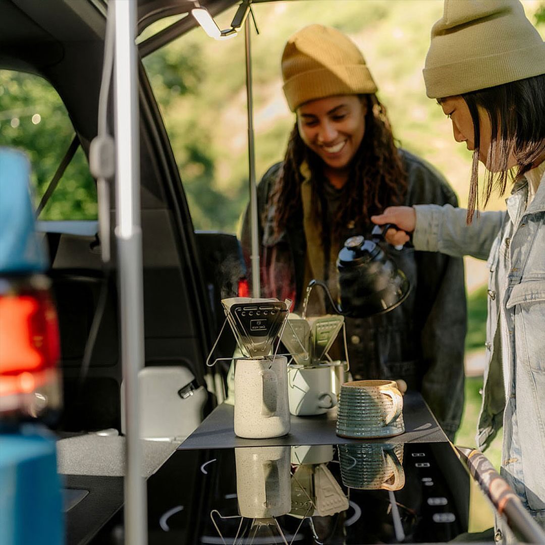 Two women prepare pour-over coffee at the back of a car in a natural setting, featuring outdoor living and vehicle-supported leisure.
