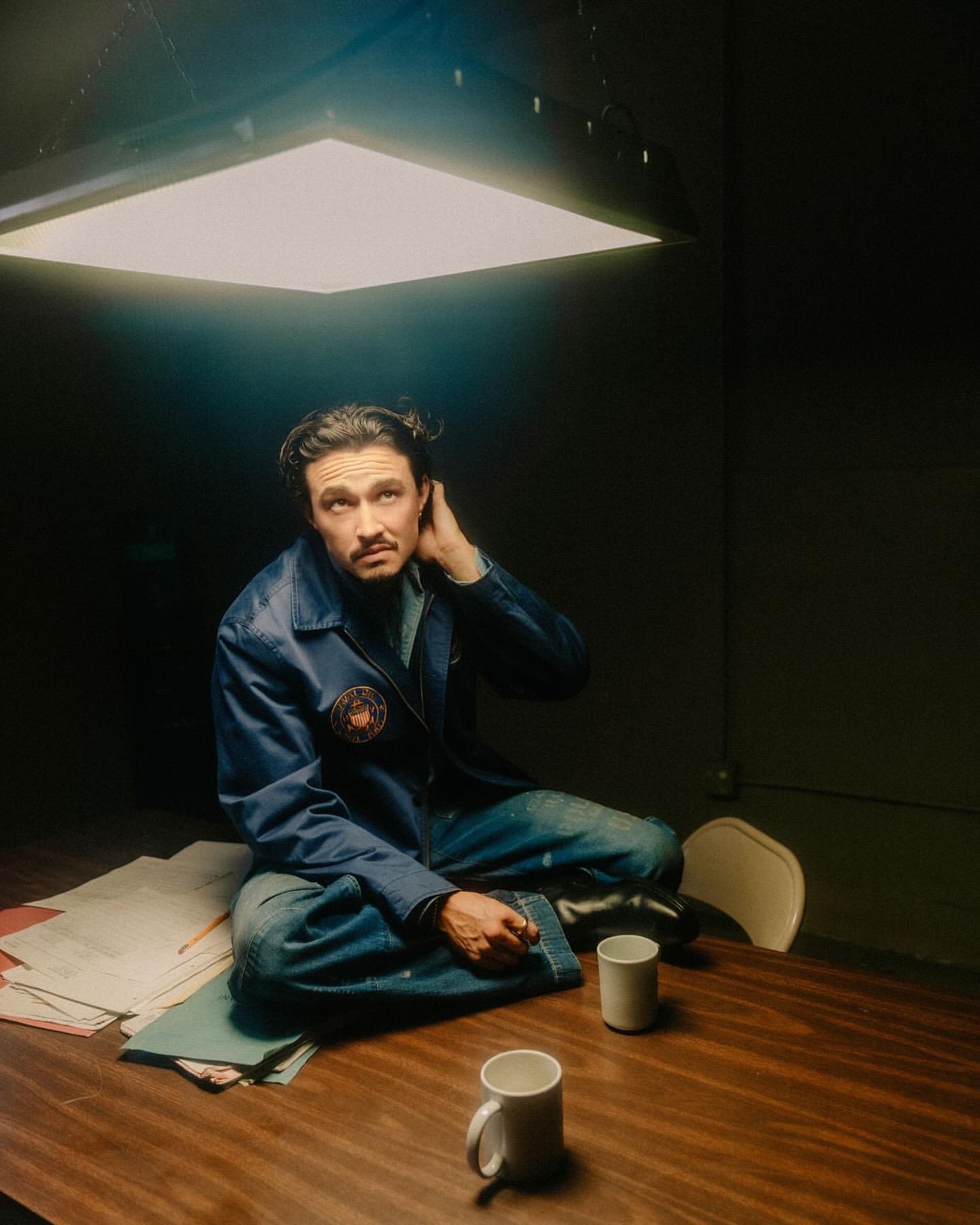 Adult male with mustache poses, seated on table with documents, under bright light in dark room. Wearing blue jacket, looking up.