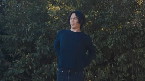 Adult male with dark hair stands outdoors in a blue sweater and jeans with a lush green background of trees and foliage in a natural setting.