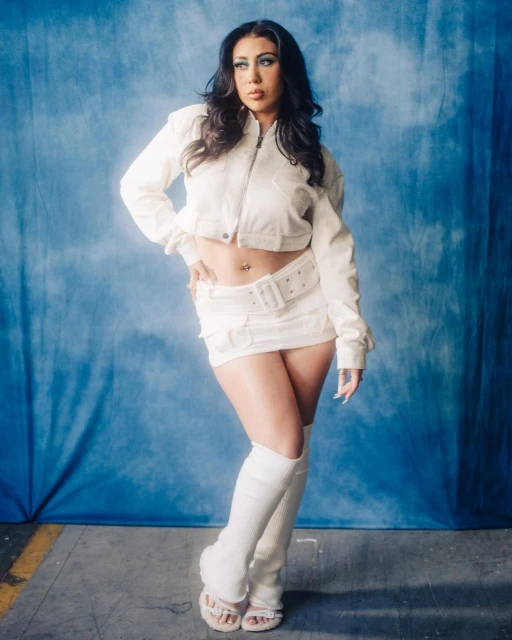 Portrait of a brunette woman in a white outfit posing against a blue cloud-like backdrop in a studio setting with soft lighting.