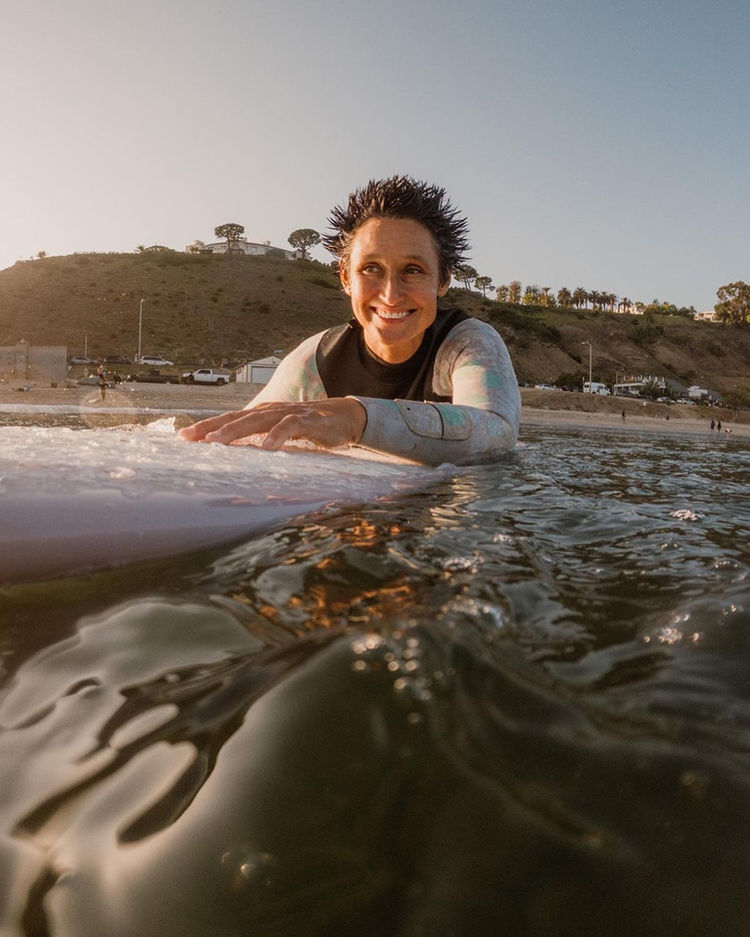 Smiling woman in wetsuit paddles on surfboard in ocean, low-angle view of water, beach, and hillside at sunrise.