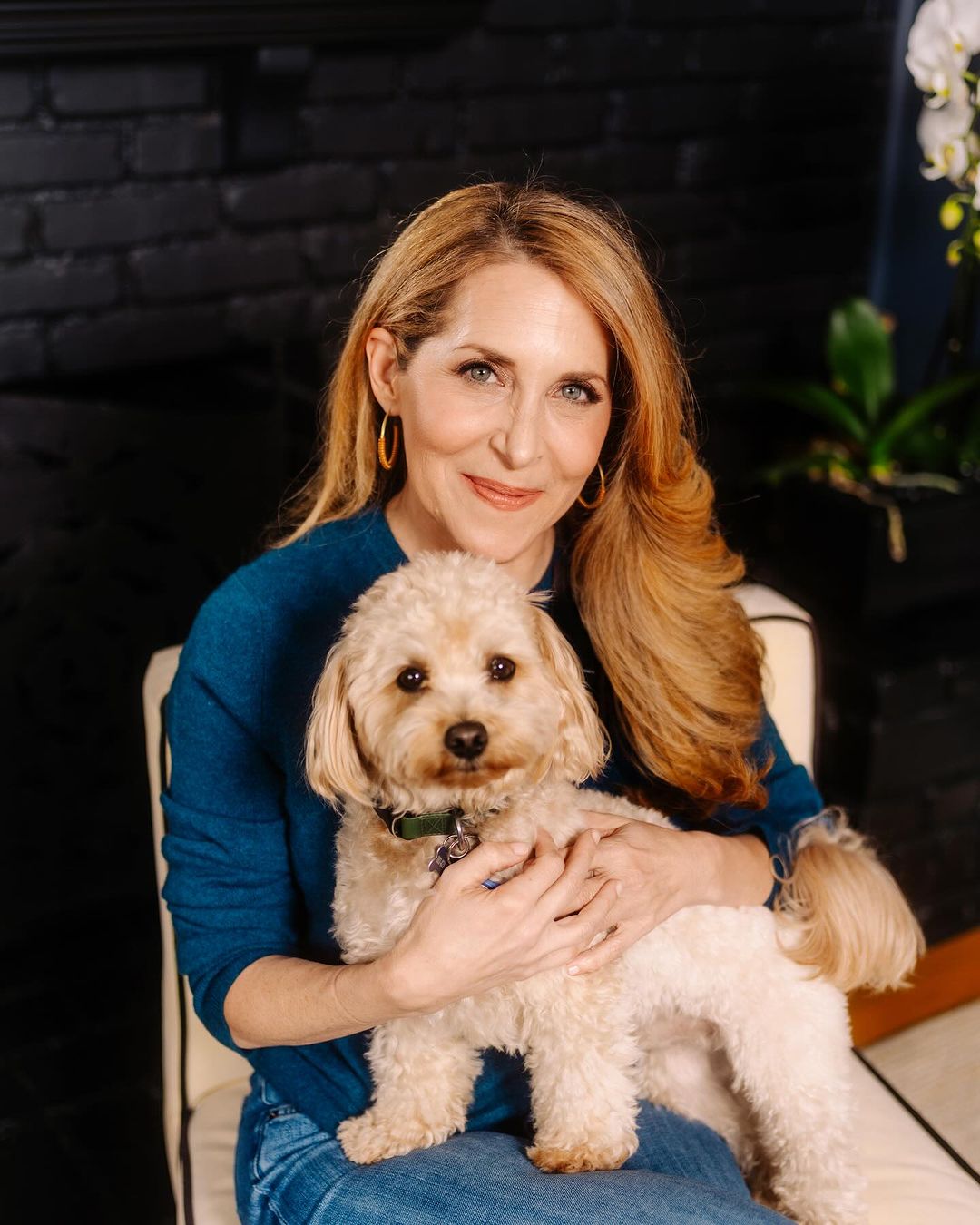 Indoor portrait of a woman with long blonde hair, wearing a blue sweater, holding a small tan-colored dog, sitting on a striped chair.