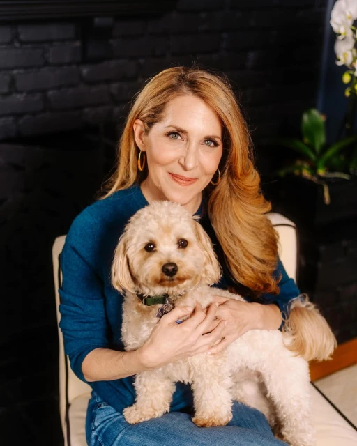 Indoor portrait of a woman with long blonde hair, wearing a blue sweater, holding a small tan-colored dog, sitting on a striped chair.