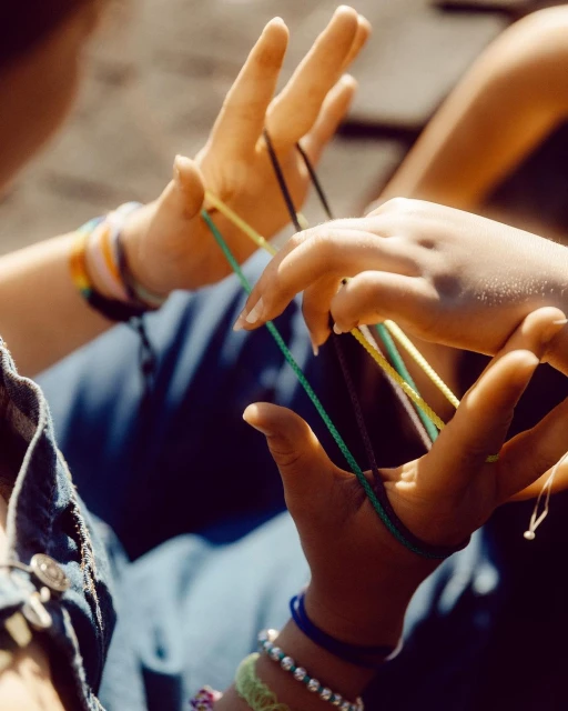 Close-up shot features a person's hands manipulating colored string to create a cat's cradle, bracelets visible on the person's wrist.