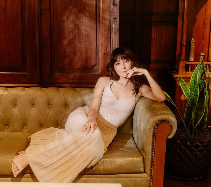 A woman with dark hair, wearing a light top and skirt, is posed on an olive-green sofa indoors with a potted plant in the background.