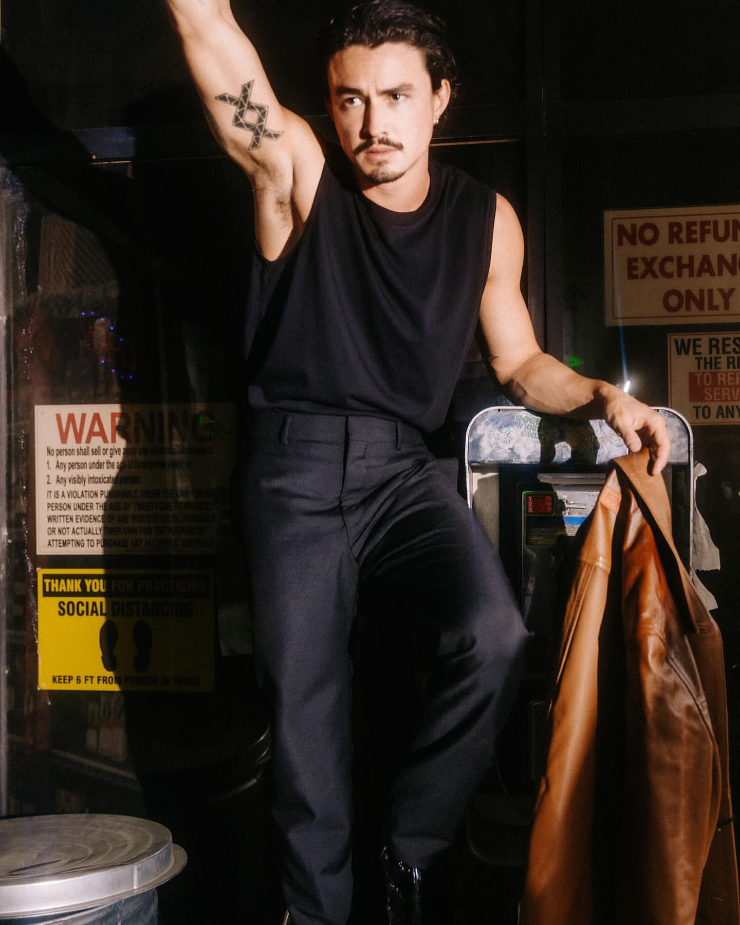 A man poses indoors wearing a black tank top and pants, an arm raised with a tattoo, holding a brown jacket in a dimly lit area.