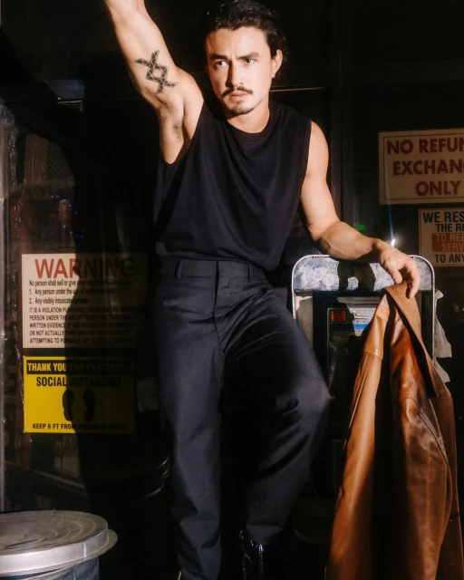 A man poses indoors wearing a black tank top and pants, an arm raised with a tattoo, holding a brown jacket in a dimly lit area.