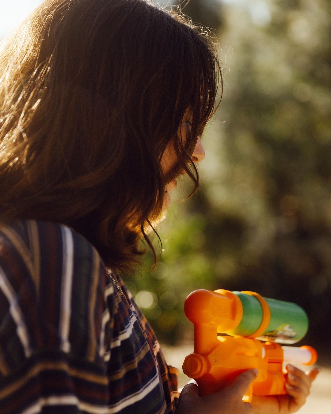 Young woman in profile holds an orange and green water gun, trees blurred in the background in an outdoor setting with natural lighting.