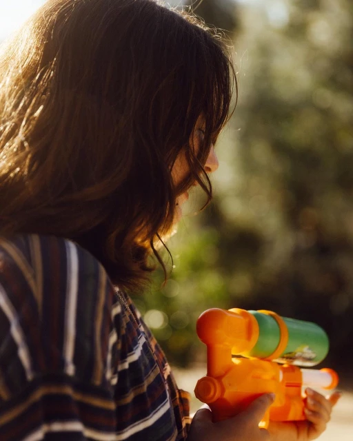 Young woman in profile holds an orange and green water gun, trees blurred in the background in an outdoor setting with natural lighting.