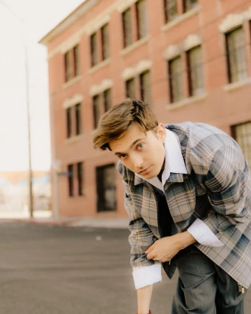 A young man in a grey plaid jacket poses in an urban outdoor setting in front of a brick building on a sunny day.