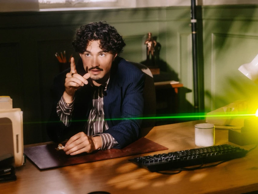 An adult male with curly brown hair sitting at a wooden desk in an office setting, wearing a striped shirt and blue blazer, indoor lighting.