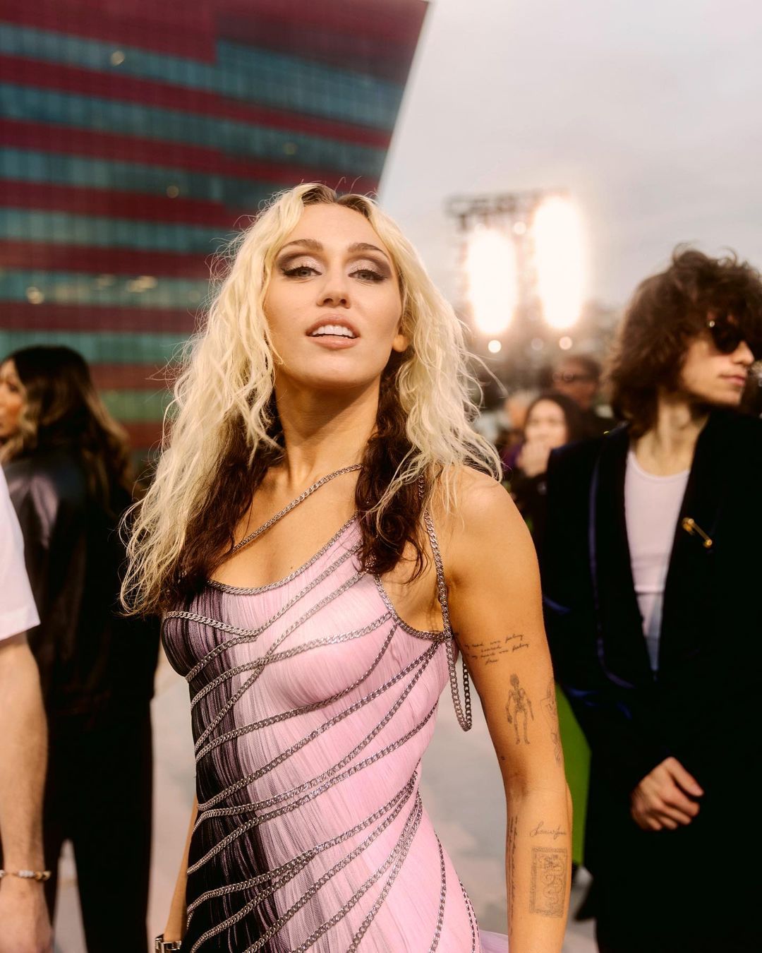 Miley Cyrus poses in a pink and black chain-detailed dress at an event. She is surrounded by people and a large building.