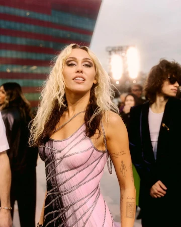 Miley Cyrus poses in a pink and black chain-detailed dress at an event. She is surrounded by people and a large building.