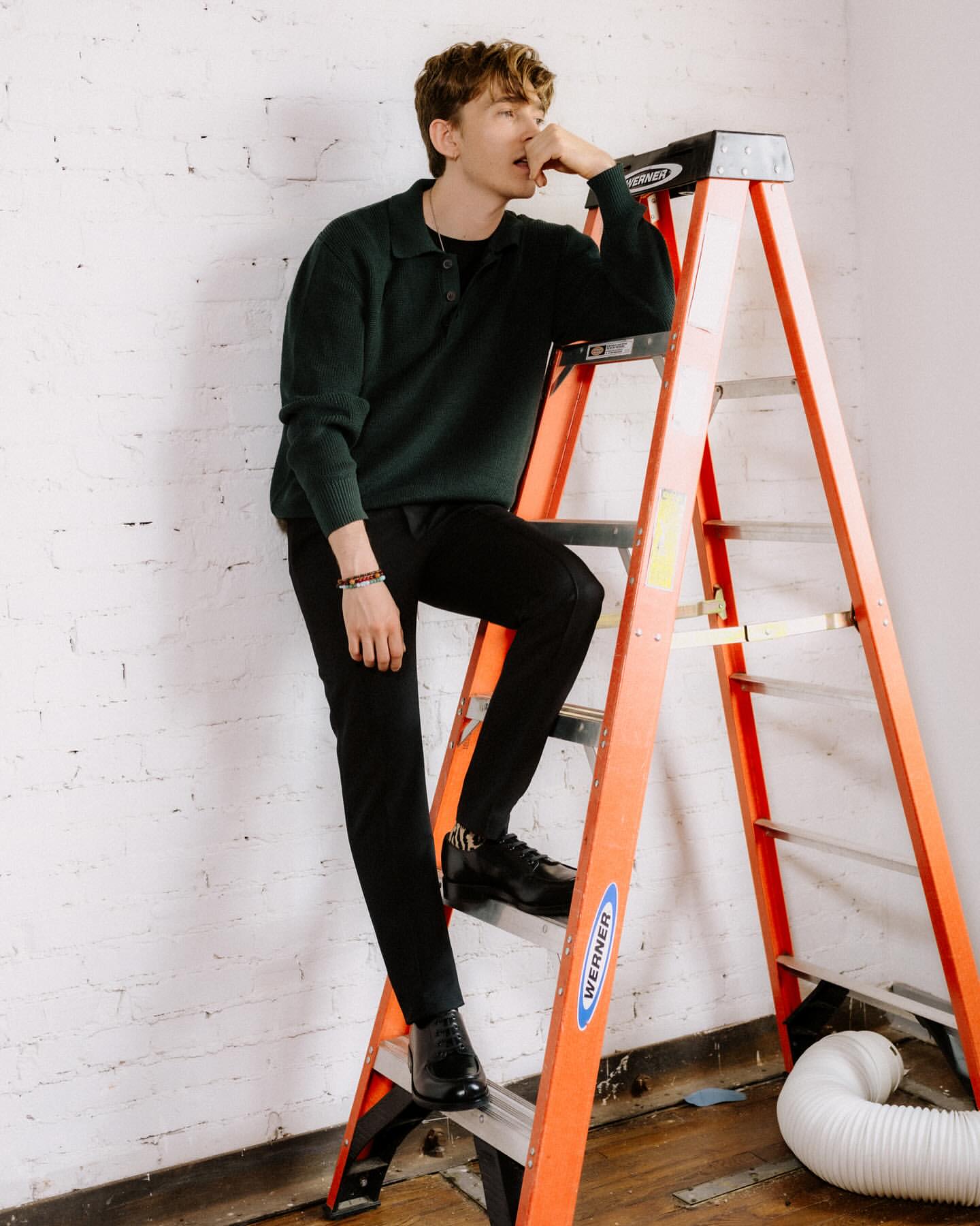 A young man in a dark sweater and black pants leans casually on an orange ladder in a photo studio, posing for a fashion-style portrait.