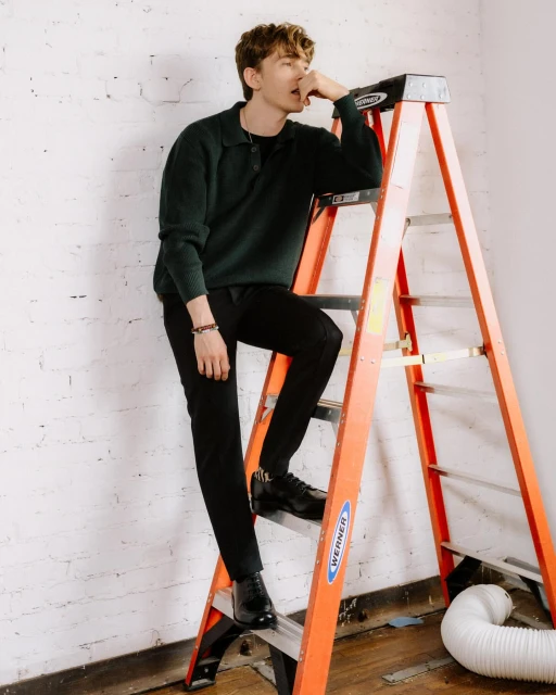 A young man in a dark sweater and black pants leans casually on an orange ladder in a photo studio, posing for a fashion-style portrait.