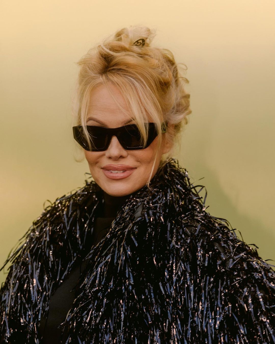 Close-up of adult female with blonde updo, sunglasses, and glittering fringed jacket, standing in front of a neutral light-yellow background.