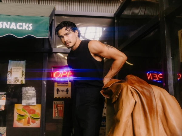 Man wearing a black tank top and dark trousers standing outdoors near a storefront with a brown jacket draped nearby.